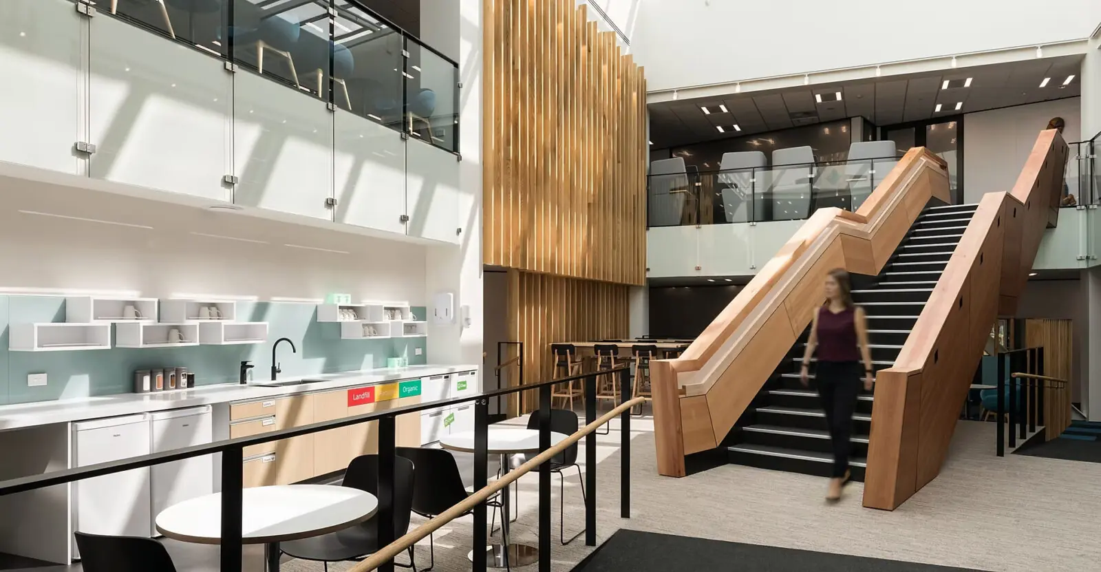 Contemporary office atrium with timber staircase, glass balustrades, café kitchenette, recycling station, vertical wood slat feature wall, skylight, and blurred person walking through a bright workplace interior.