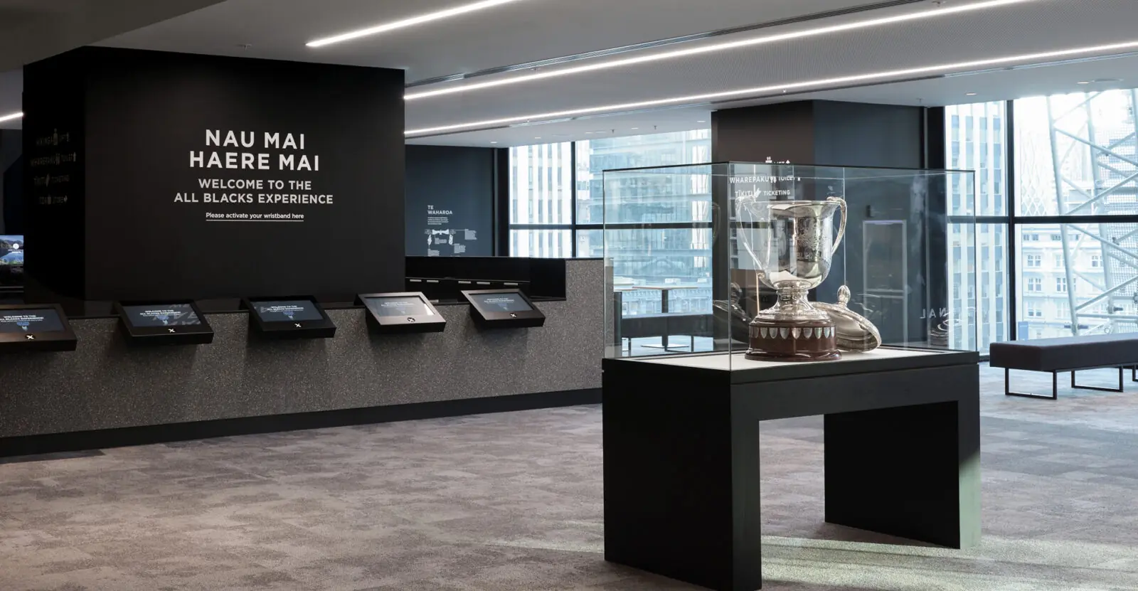 All Blacks Experience museum entrance with interactive screens, trophy display case, modern black interior, and wall text reading “Nau Mai Haere Mai Welcome to the All Blacks Experience.”