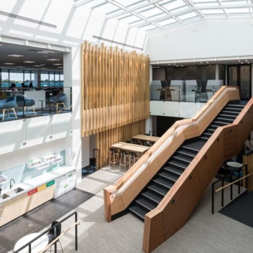 Bright office atrium with skylight ceiling, timber feature wall, glass balustrades, wooden staircase, café seating, kitchenette, recycling bins, and contemporary workplace interior design.