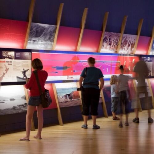 Museum exhibition gallery with visitors viewing a colorful historical display wall, archival photos, illustrated panels, artifacts, and text including “Assault from the shore.”