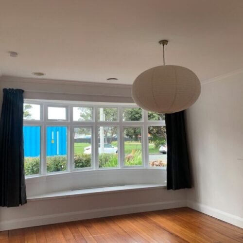 Empty renovated living room with polished timber floors, large bay window, black curtains, white walls, built-in window bench, and round paper pendant light.