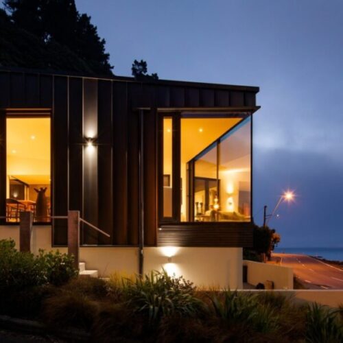 Contemporary black-clad hillside home at dusk with warm interior lighting, large corner windows, coastal road view, landscaped garden, and modern architectural design.