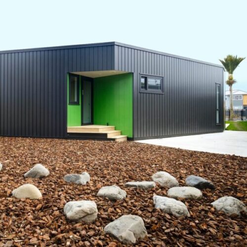Modern black metal-clad modular building with bright green recessed entrance, wooden steps, rock landscaping, bark mulch, and contemporary exterior architecture.