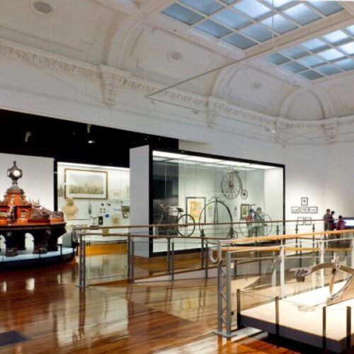 Historic museum gallery interior with polished wooden floors, ornate ceiling, glass display cases, antique bicycles, archival objects, and heritage exhibition design.
