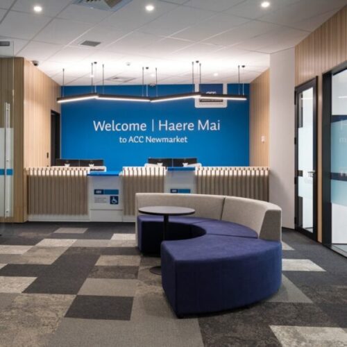 ACC Newmarket reception area with blue “Welcome | Haere Mai to ACC Newmarket” feature wall, timber slat front counters, accessible service desk, curved blue lounge seating, and patterned carpet tiles.