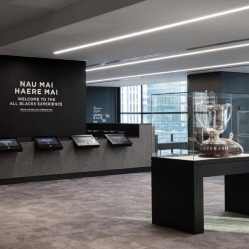 All Blacks Experience museum entrance with interactive screens, trophy display case, modern black interior, and wall text reading “Nau Mai Haere Mai Welcome to the All Blacks Experience.”