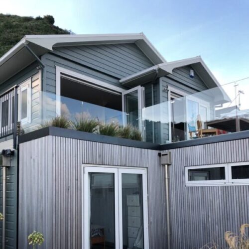 Modern coastal house exterior with glass balcony balustrades, grey weatherboard cladding, timber lower facade, outdoor seating, and large sliding doors.