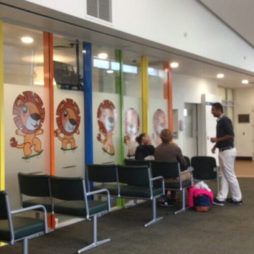 Children’s clinic waiting area with black chairs, glass wall decals of cartoon lions on skateboards, colourful vertical panels, and families speaking with a staff member.