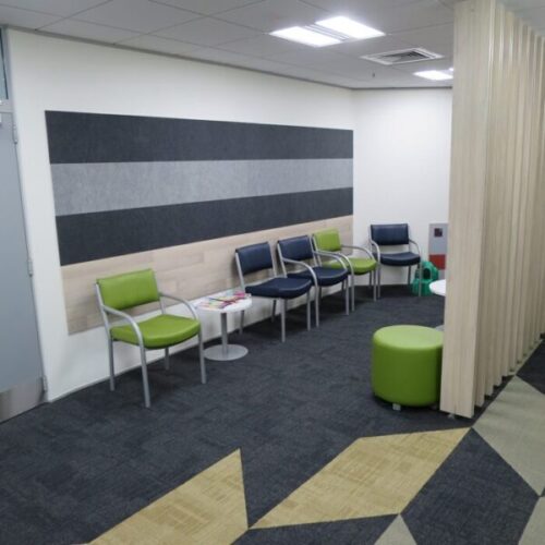 Modern healthcare waiting room with green and black chairs, striped acoustic wall panel, round side tables, patterned carpet, and pale wood privacy screen.