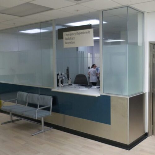 Hospital reception desk for “Emergency Department Radiology Reception,” with frosted glass partitions, bench seating, clinical lighting, and a medical staff member behind the counter.