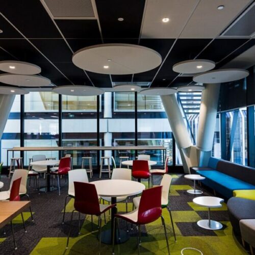 Contemporary workplace breakout area with round white tables, red and white chairs, blue sofa seating, circular acoustic ceiling panels, black ceiling grid, and large atrium windows.