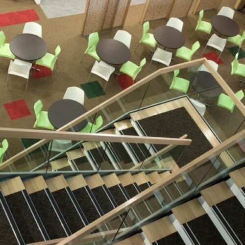 Overhead view of a public cafeteria with glass balustrade stairs, round dark tables, green and white chairs, patterned carpet squares, and light timber railings.