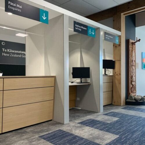 Government customer service area with “Pātai Mai Enquiries” and “Āwhina Whaiaro Self Help” signs, self-service computer stations, timber counters, blue carpet tiles, and carved Māori artwork.