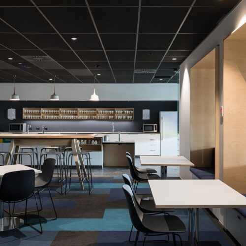 Modern office kitchen and staff breakout area with black ceiling tiles, pendant lights, long timber island bench, bar stools, open cup shelving, white cabinetry, microwaves, fridge, booth seating, black chairs, white tables, and blue geometric carpet tiles.