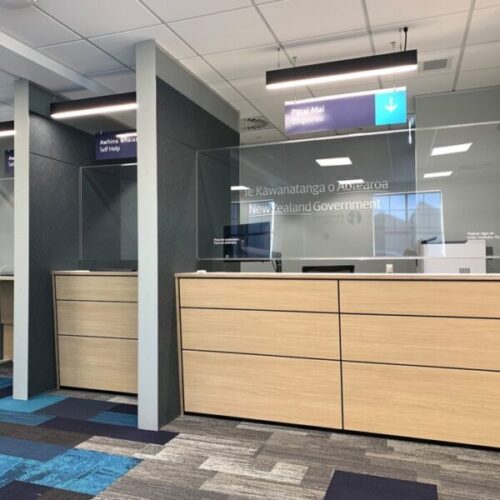 New Zealand Government service centre with wood-front enquiry counters, glass safety screens, overhead signage reading “Pātai Mai Enquiries” and “Āwhina Whaiaro Self Help,” and blue patterned carpet.