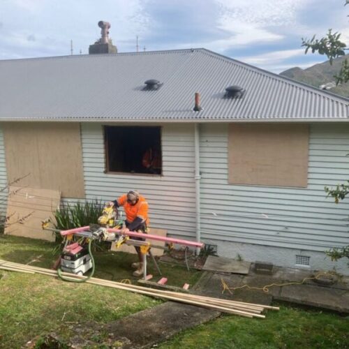 Residential renovation in progress on a weatherboard house with boarded windows, grey metal roof, exterior cladding repairs, construction tools, timber planks, and a worker using a miter saw.