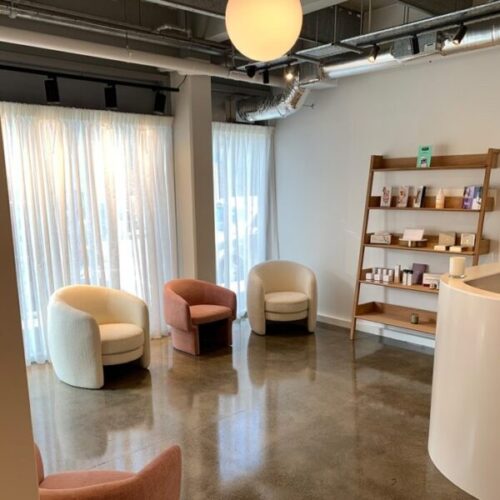 Contemporary wellness clinic waiting room with curved upholstered chairs, sheer curtains, polished concrete floor, wood retail shelving, exposed ceiling ducts, and warm pendant lighting.