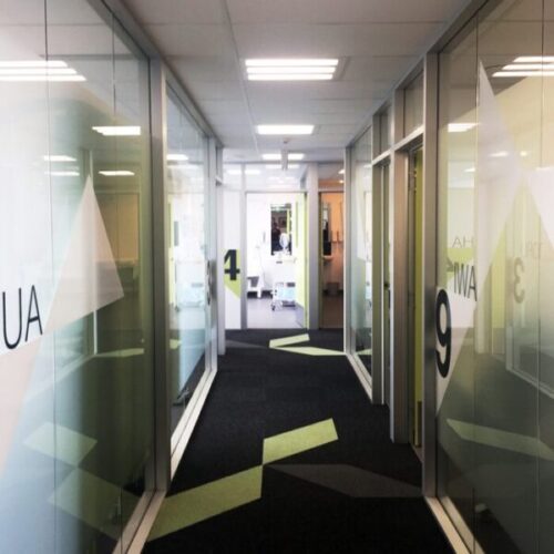 Medical clinic corridor with glass-walled rooms, numbered wayfinding graphics, dark carpet, geometric floor accents, and bright overhead fluorescent lighting.