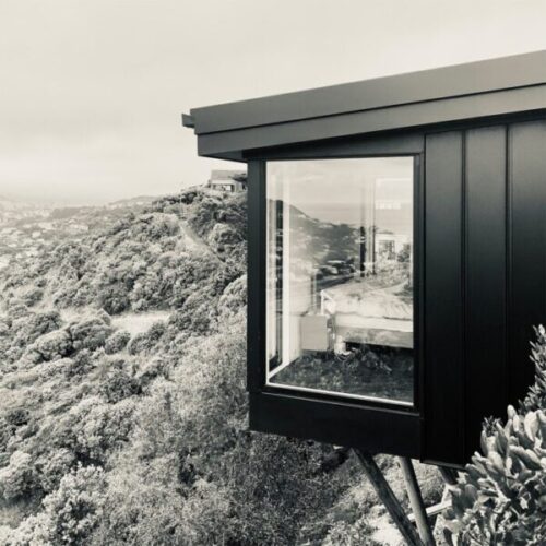 Black-and-white hillside home exterior with black metal cladding, large picture window, elevated bedroom view, coastal landscape, and rugged bush-covered surroundings.