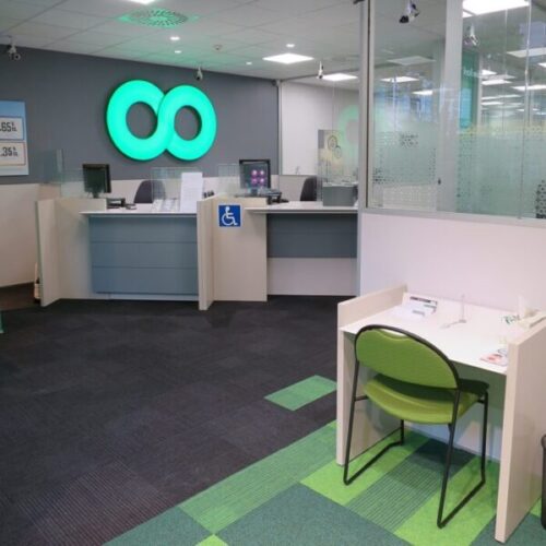 Cooperative Bank branch interior with illuminated green infinity logo, accessible service counter, glass partitions, teller desks, green accent carpet tiles, and customer consultation table.