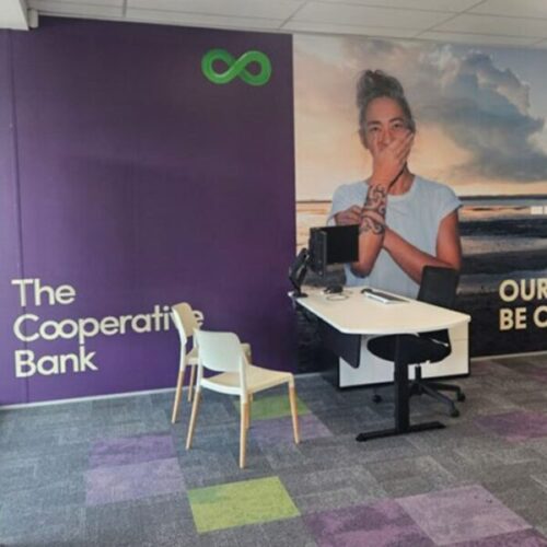 Cooperative Bank customer consultation area with purple branded wall reading “The Cooperative Bank,” workstation desk, white visitor chair, portrait mural, green infinity logo, and patterned carpet tiles.