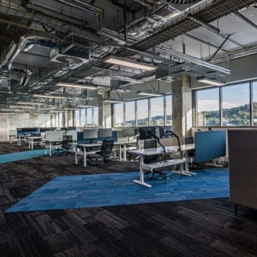 Industrial open-plan office with exposed ductwork, cable trays, suspended linear lights, workstations, monitor arms, large windows, blue accent carpet zones, and panoramic hillside views.
